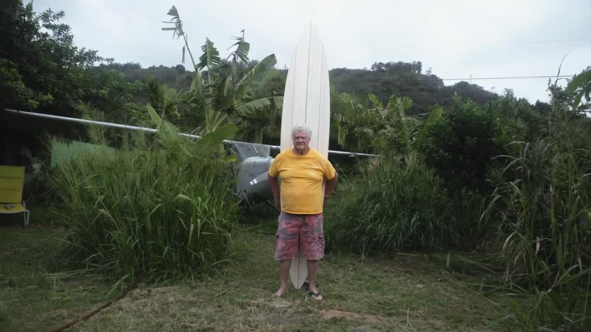 A man holding a surfboard with trees the sky and clouds in the background