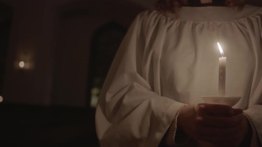 A person wearing a frock carries a candle in a dimly lit church