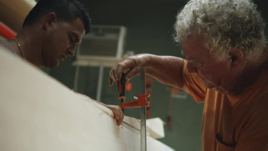 A man holds pieces of wood together as another man screws a tool in to the wood
