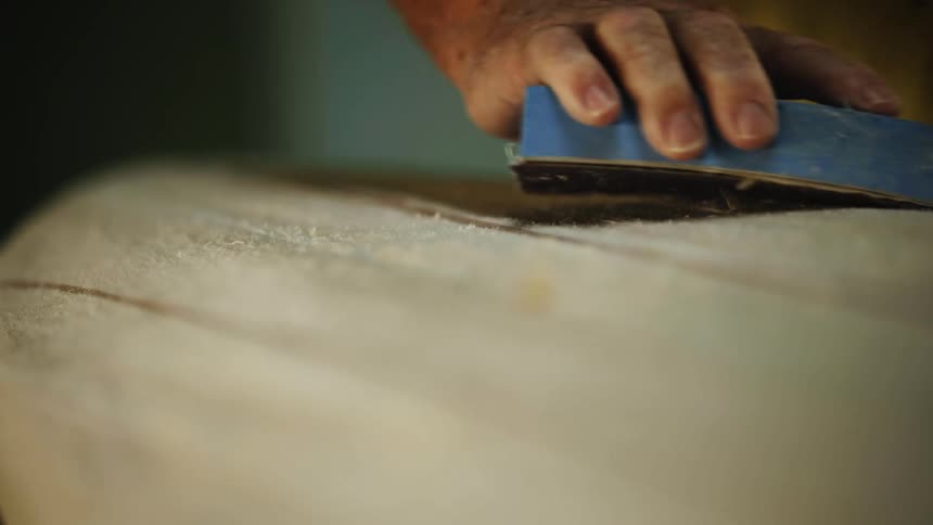 A man hand sands and smooths a surfboard