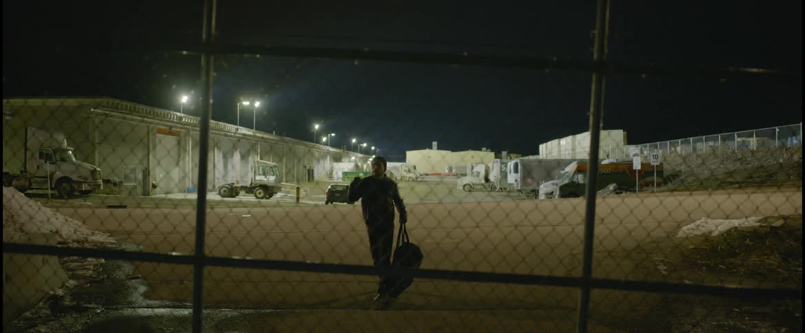 A man carries a duffle bag through the parking lot of a depot and attempts to throw it over a chain link fence
