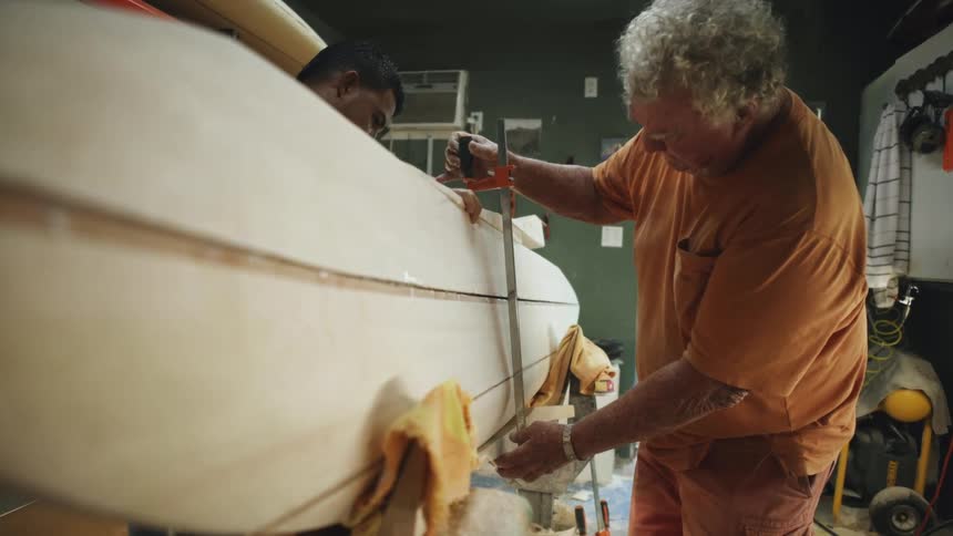 Two men in a room using a tool on a surfboard with a wall in the background