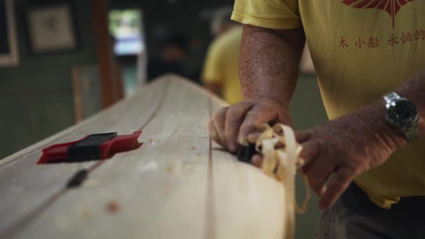 A man shaves wood from a surfboard using a hand plane