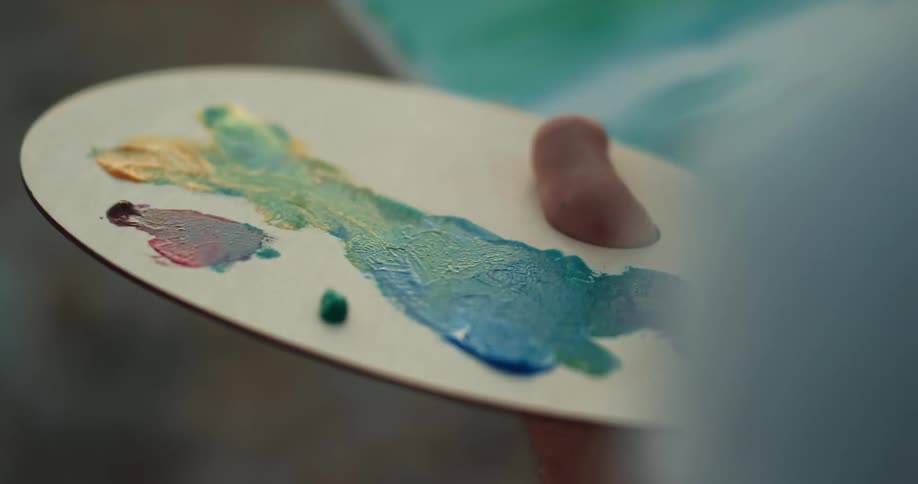 A person holding a paint palette while painting on a canvas with a brush