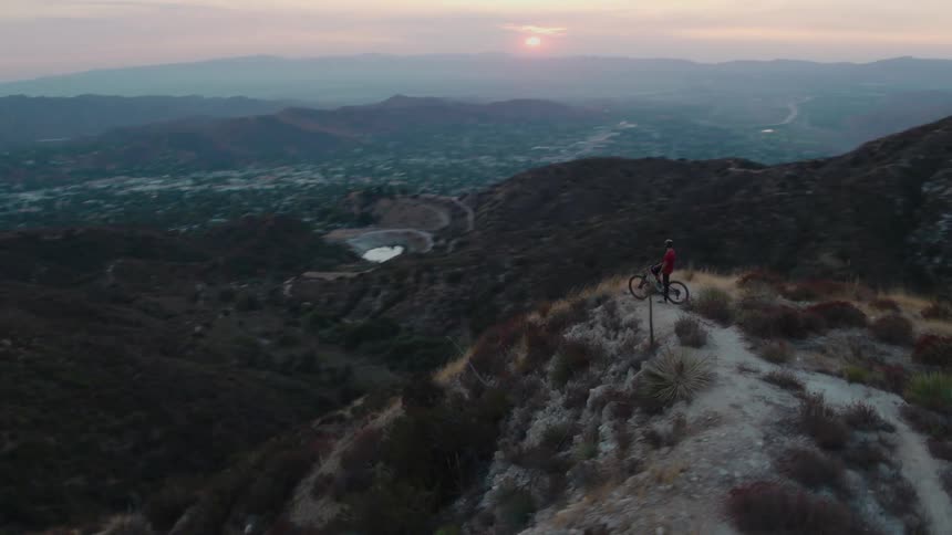 Person stands next to bicycle on trail on top of narrow hill looking toward town and with sun sitting low in red tinted sky