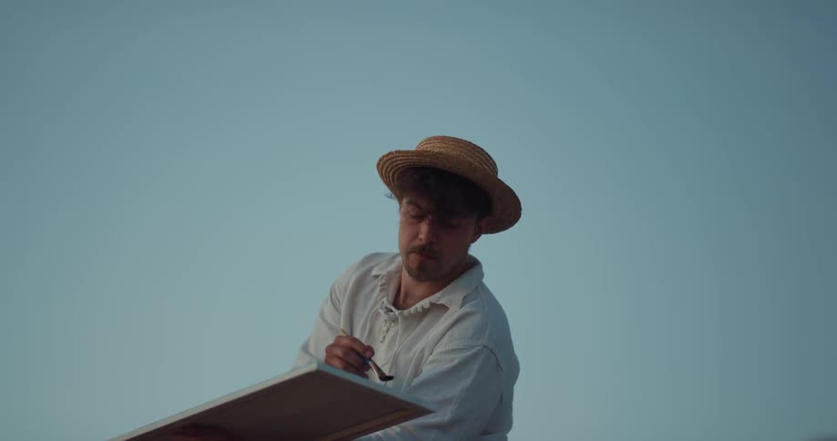 Man wearing a straw hat holds a canvas as he paints it with a paint brush under a blue sky
