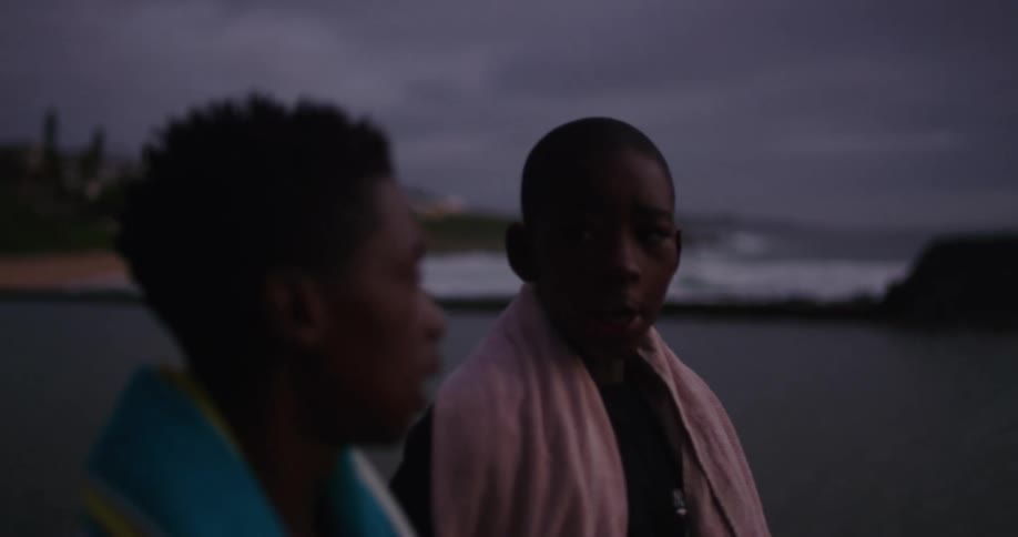Two boys with towels walking on a beach with the ocean sky and clouds in the background