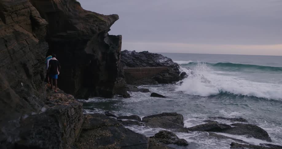 Two men in shorts walk along the edge of a large rock near the ocean