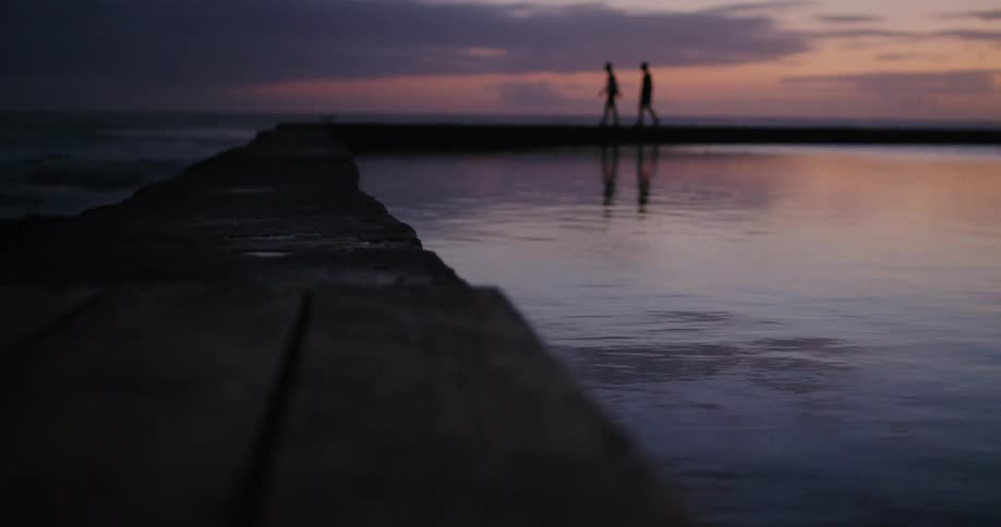 Two people walk along stone man made causeway silhouetted against red sky as waves crash and water ripples