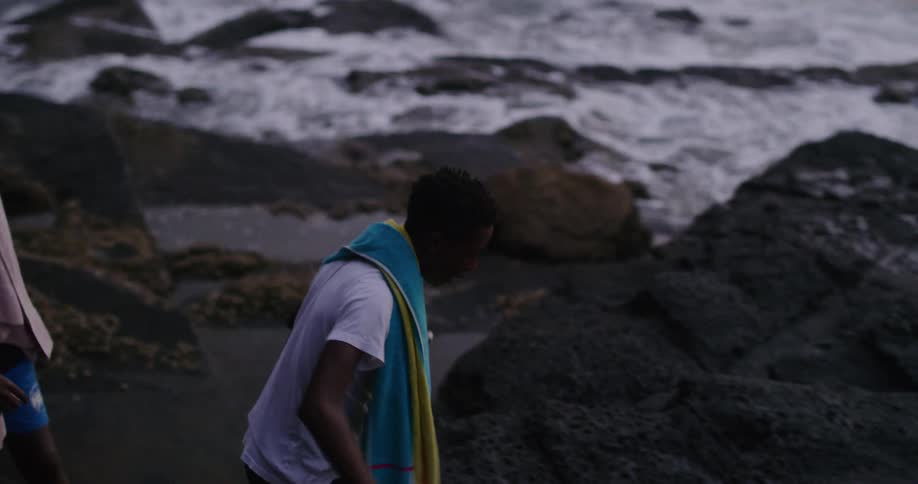 Two front walk on large boulders along the coast with towels draped over their shoulders