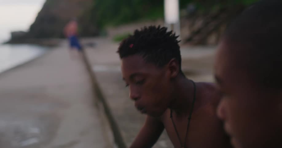 A boys sits on the shore and instructs another boy how to move his arms and breathe while swimming