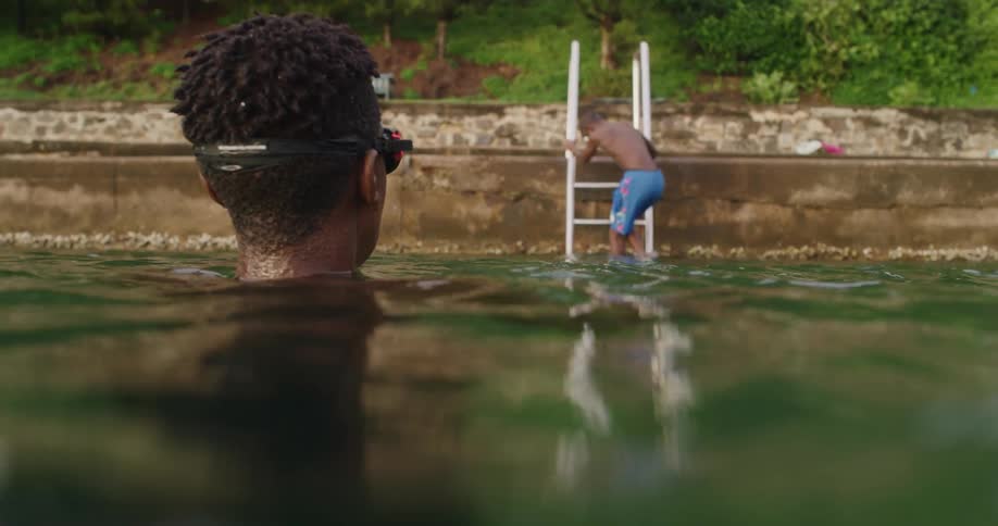 A boy slowly climbs down a ladder into the water where his friend is treading water