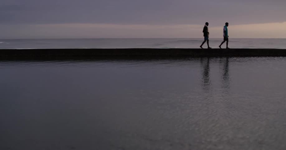 Two men with towels around their necks and wearing shorts walk on a pier in the ocean