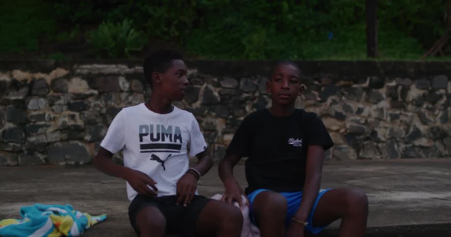 A pair of boys carrying towels around their necks and wearing swim trunks sit down and begin to take off their shirts