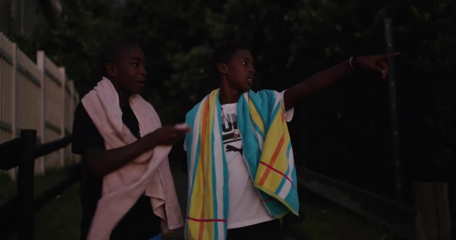 Two black men with towels around their necks smile and point while standing near the forest