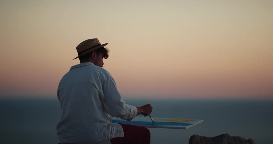 A man with a hat painting on a canvas with a paintbrush with the sky and clouds in the background