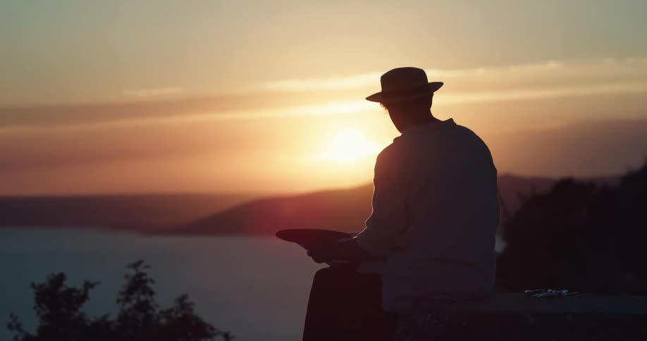 A silhouette of a person with a hat painting with a paintbrush with the sky and clouds in the background