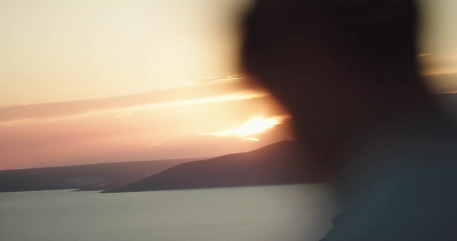 Young man wearing a straw hat looks at the sunset over the mountains near the ocean