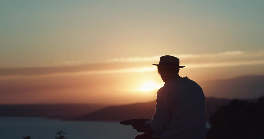 Silhouette of a man wearing a hat and painting a picture with a bright colorful sunset on the horizon