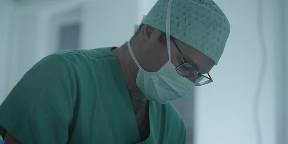A doctor wearing cap mask and scrubs looks down while working on a patient