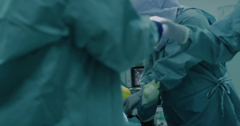 Medical personnel wearing scrubs and protective gear move around patient in operating theater