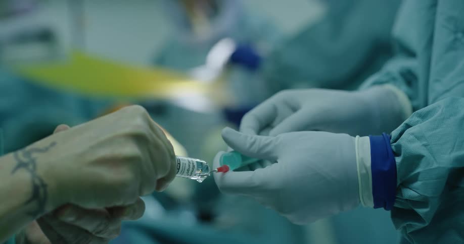 A gowned and gloved doctor fills a syringe with medicine from a tube held by tattooed hands