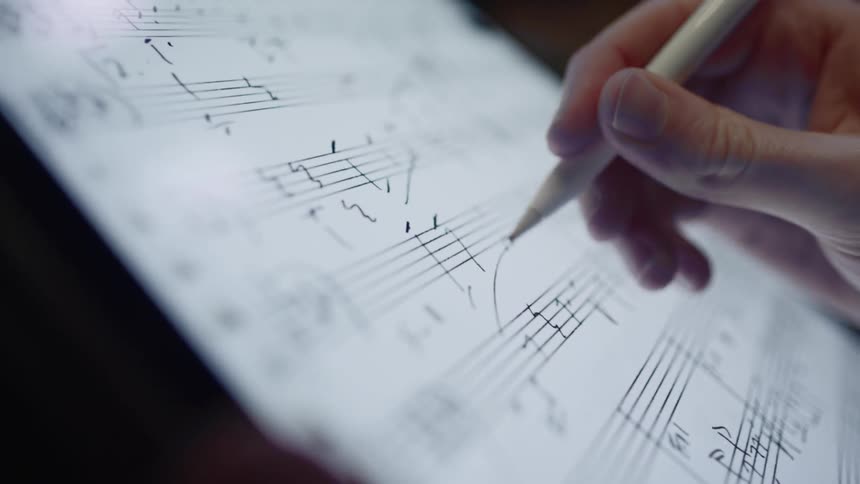 A hand holding a stylus and tablet draws notes on sheet music