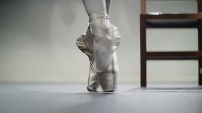 A ballerina moves sideways on the tips of her toes in front of a chair in a large room