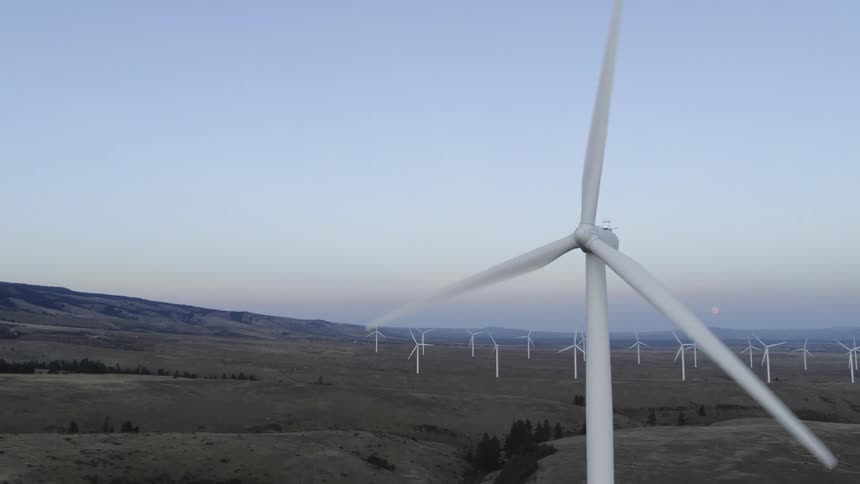 Pullback from a low angle closeup of a wind turbine to reveal an entire wind farm with the moon hanging low above the horizon