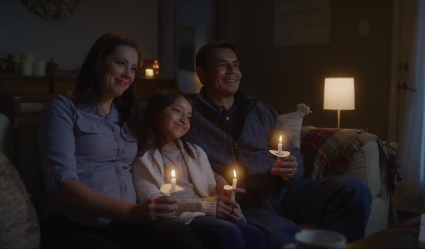 A man woman and child sit on the couch at home holding candles as the man reaches his arm and puts around his child and the woman