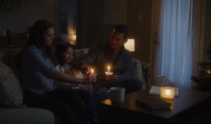 A man uses a lighter to light candles being held by his family as they sit together on a sofa in the living room of a house at night