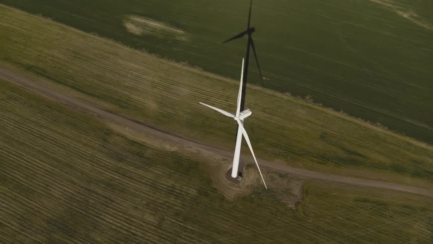 A wind turbine casts a long shadow as it spins above green farm fields