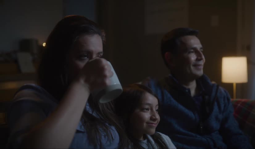 A little girl sits on the couch between her mom and dad as she holds a book and smiles and her mom drinks out of a mug