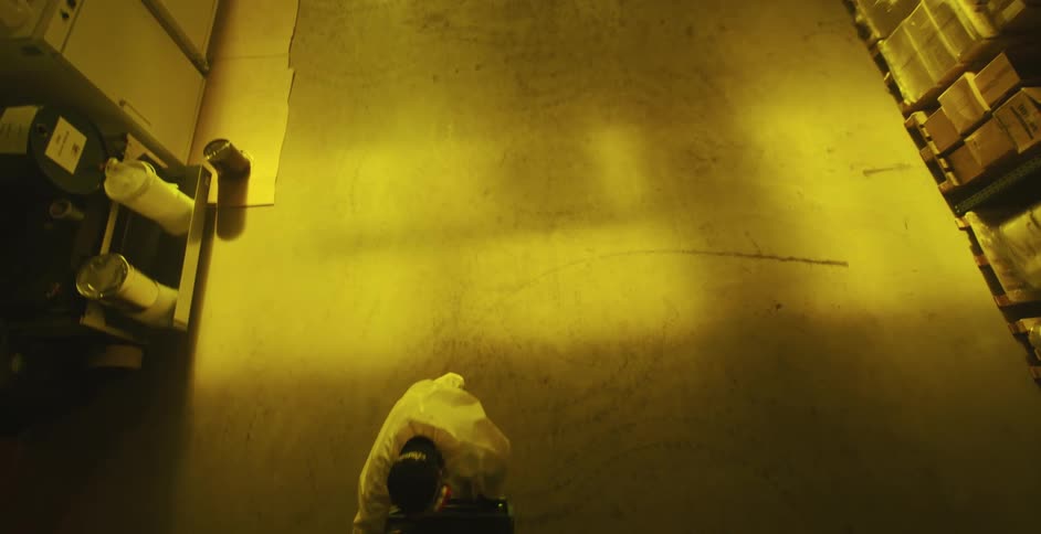 A worker in a hazmat suit wheels a cart of cylinders down a row in a warehouse
