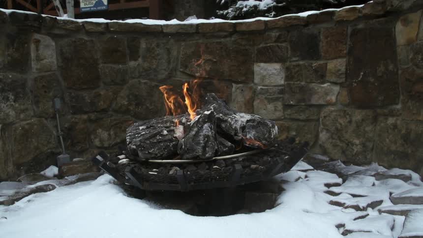 Logs burn in an outdoor fire pit, as snow falls gently