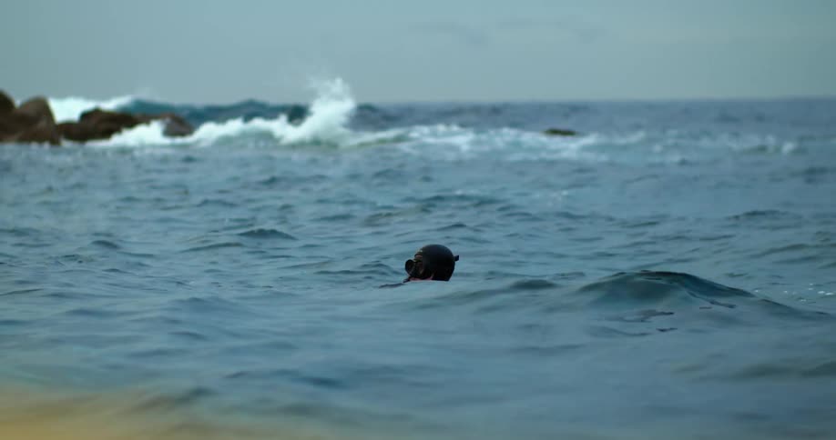 Waves are crashing into rocks as someone wearing a mask and snorkel rises from the water