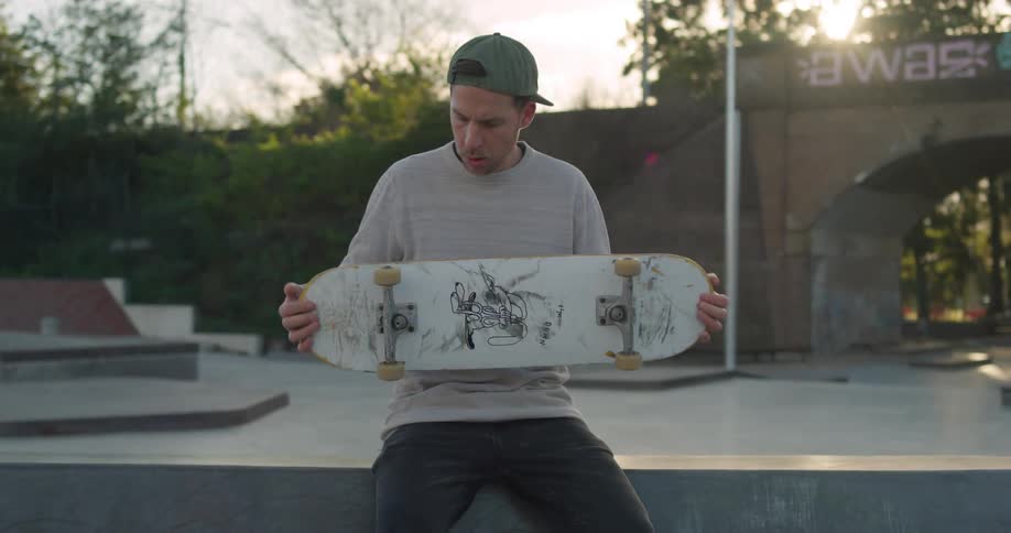 Young man adjusting the wheels of a skateboard sitting in a park at sunset