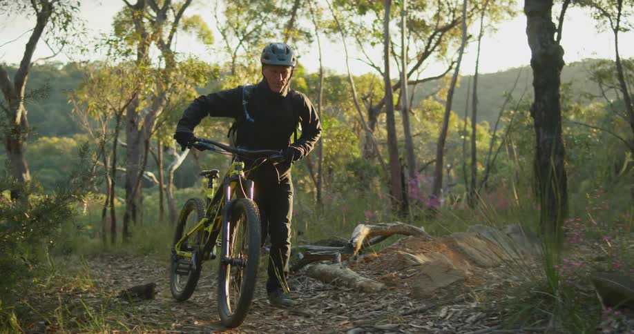 A man is walking his bicycle through a forest at dusk