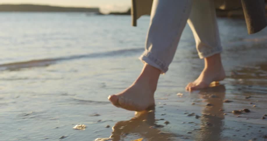 A person walking on a sandy beach with waves washing up at their feet with the sky and clouds in the background