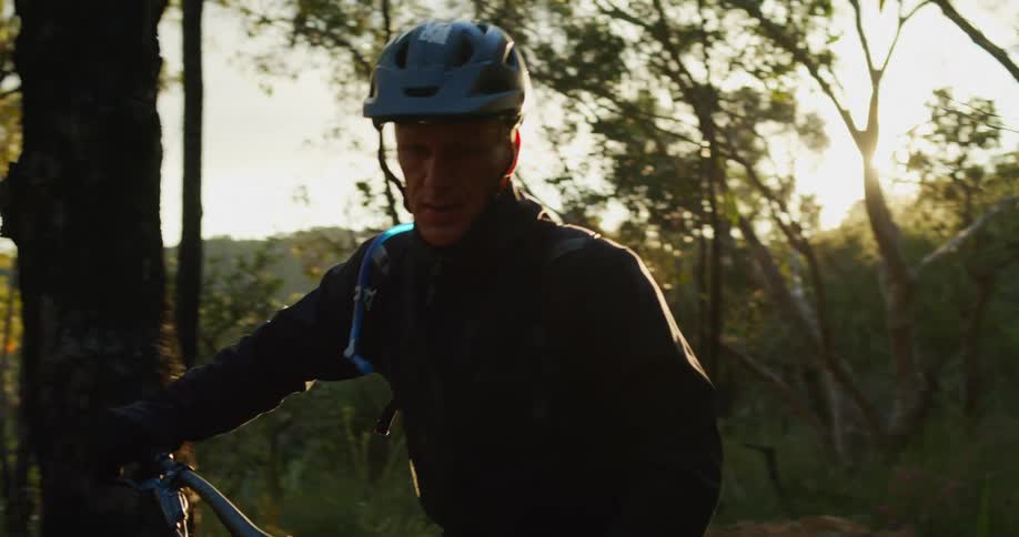 A man is walking his bike through a wooded area at dusk