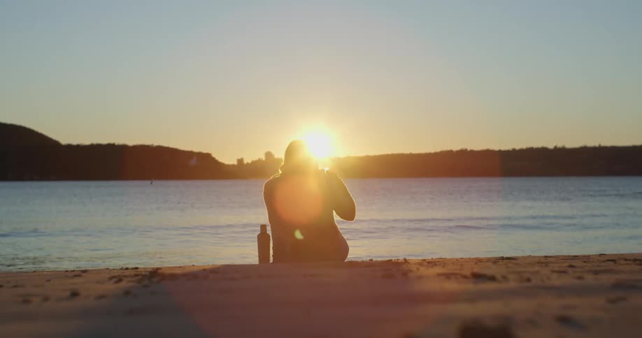 A woman sits on a lakeside beach with a drink container beside her