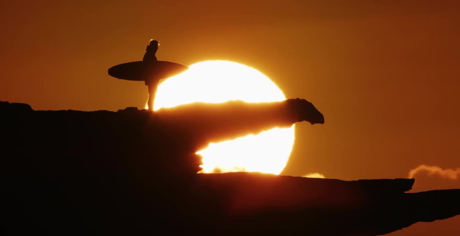Silhouette of a man holding a surfboard standing on a promontory in front of the sun at sunset