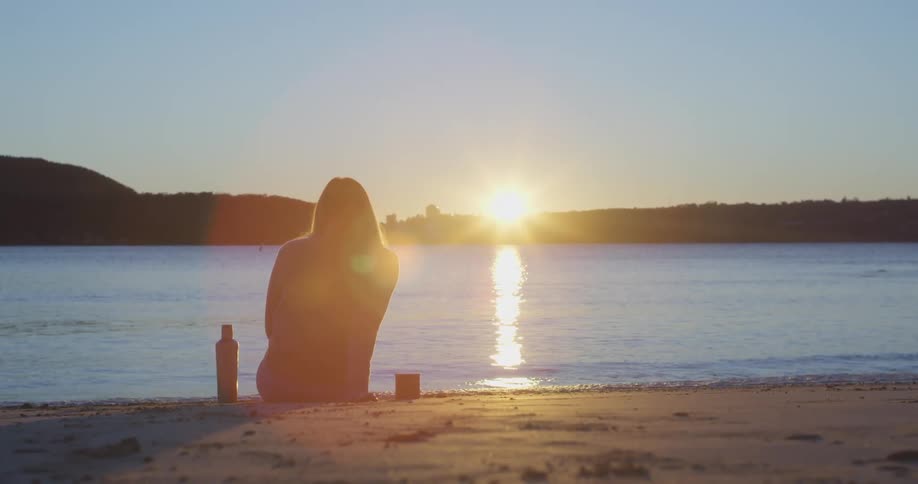 Silhouette of a young woman sitting on the sand near a lake at sunset
