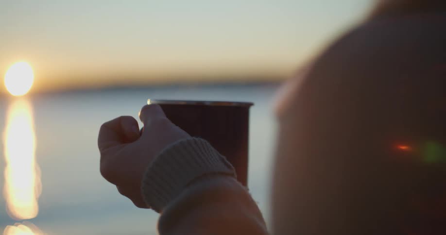 A person relaxes on the coast at sunset with a cup of steaming coffee