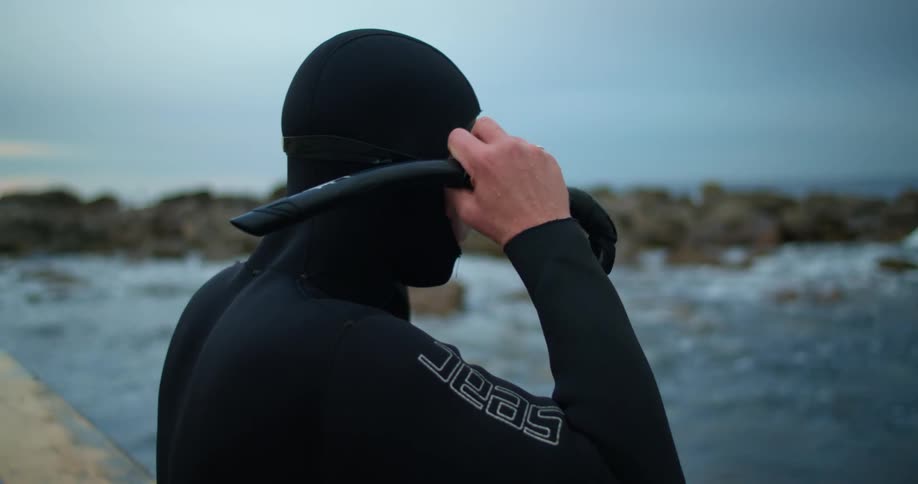 A diver sitting next to the ocean puts on his diving goggles and snorkel