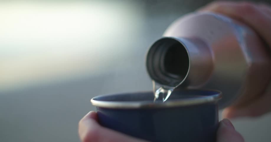 Hands hold a flask and cup as hot water pours into the cup