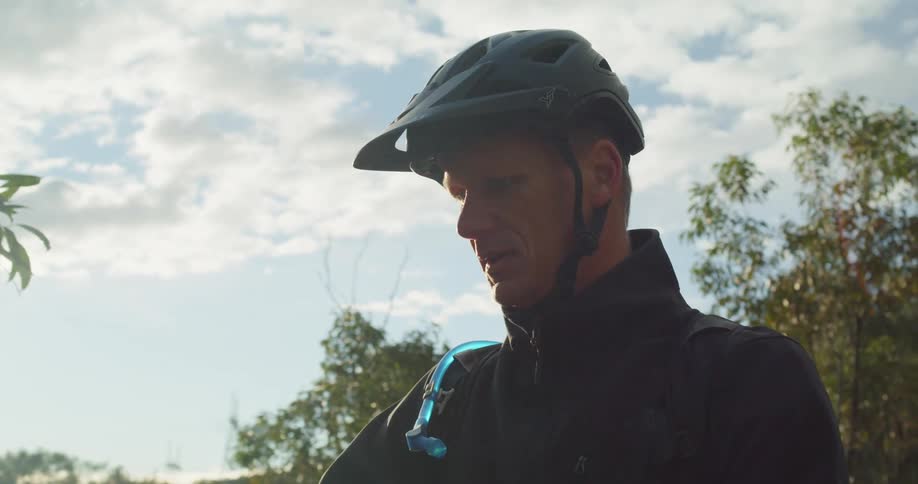 Man wearing bicycle helmet stands outdoors in woods on sunny day considering