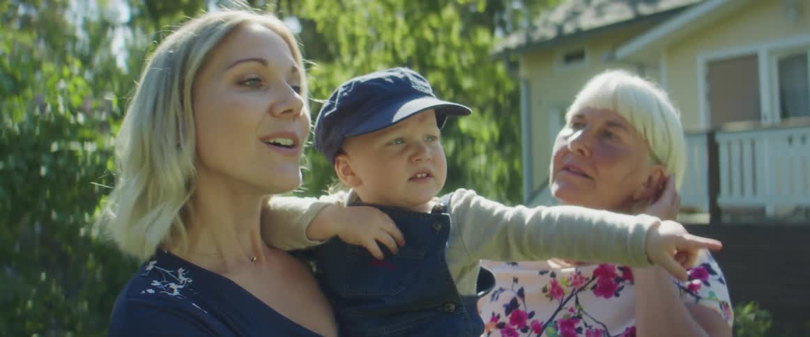 A woman holding a baby boy while talking to another woman with a house trees the sky and clouds in the background