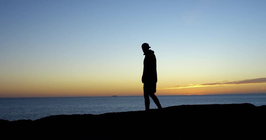 A man walks across a rocky coastline before turning to face the ocean at sunset