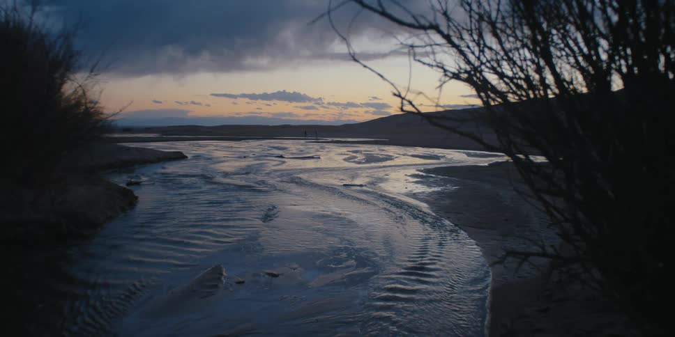 Shallow water ripples as it flows over sand near shore and brush at sunrise